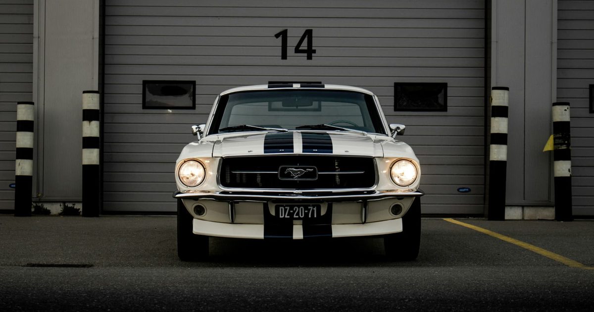 White classic Ford Mustang with dark racing stripes parked in front of a garage door